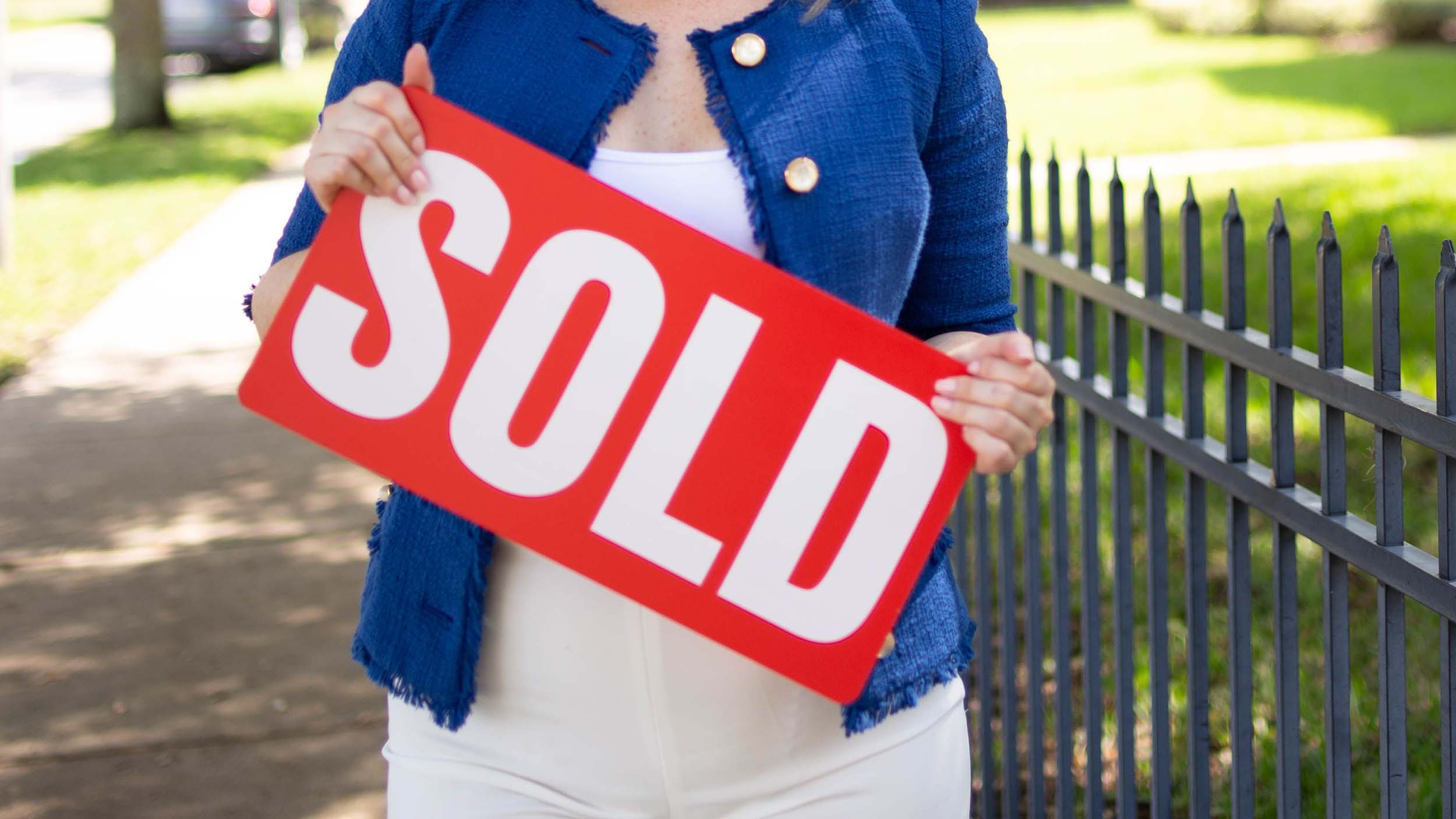 Woman holding a red sold sign beside a fence in a residential street setting