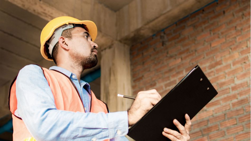 Construction inspector in hard hat and safety vest writing notes on a clipboard inside a building under construction